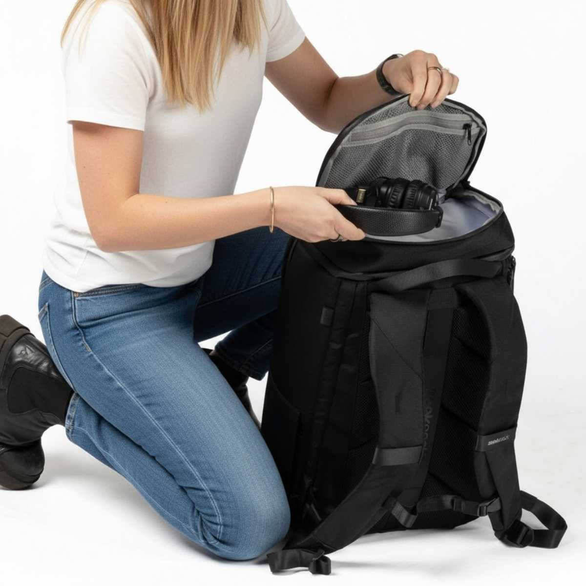 Kobieta w białej koszulce i dżinsach otwiera górną kieszeń czarnego plecaka Zeeksack, studio na białym tle.