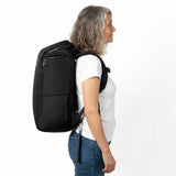 Kobieta w średnim wieku w białej koszulce i dżinsach prezentuje plecak Zeeksack w profilu, studio na białym tle.