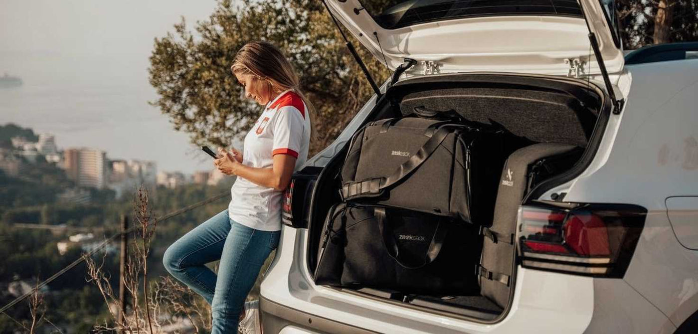 Kobieta w dżinsach i koszulce w stylu narodowym opiera się o samochód z otwartym bagażnikiem. W środku znajdują się czarne torby podróżne Zeeksack. Kobieta patrzy na telefon, a w tle widać panoramę miasta i morze.